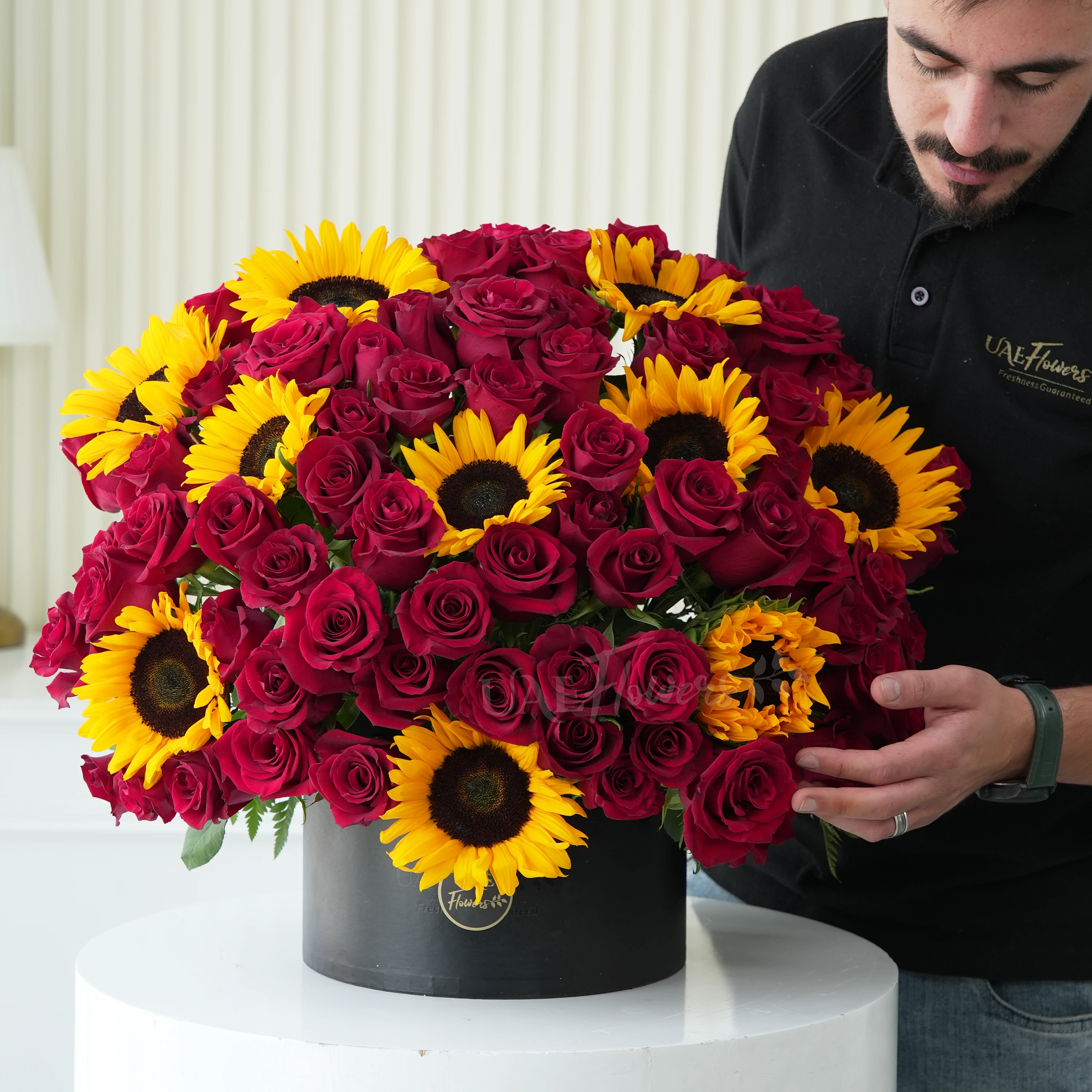 One side round arrangement of 10 sunflowers and 60 red roses with greens in a black round box.