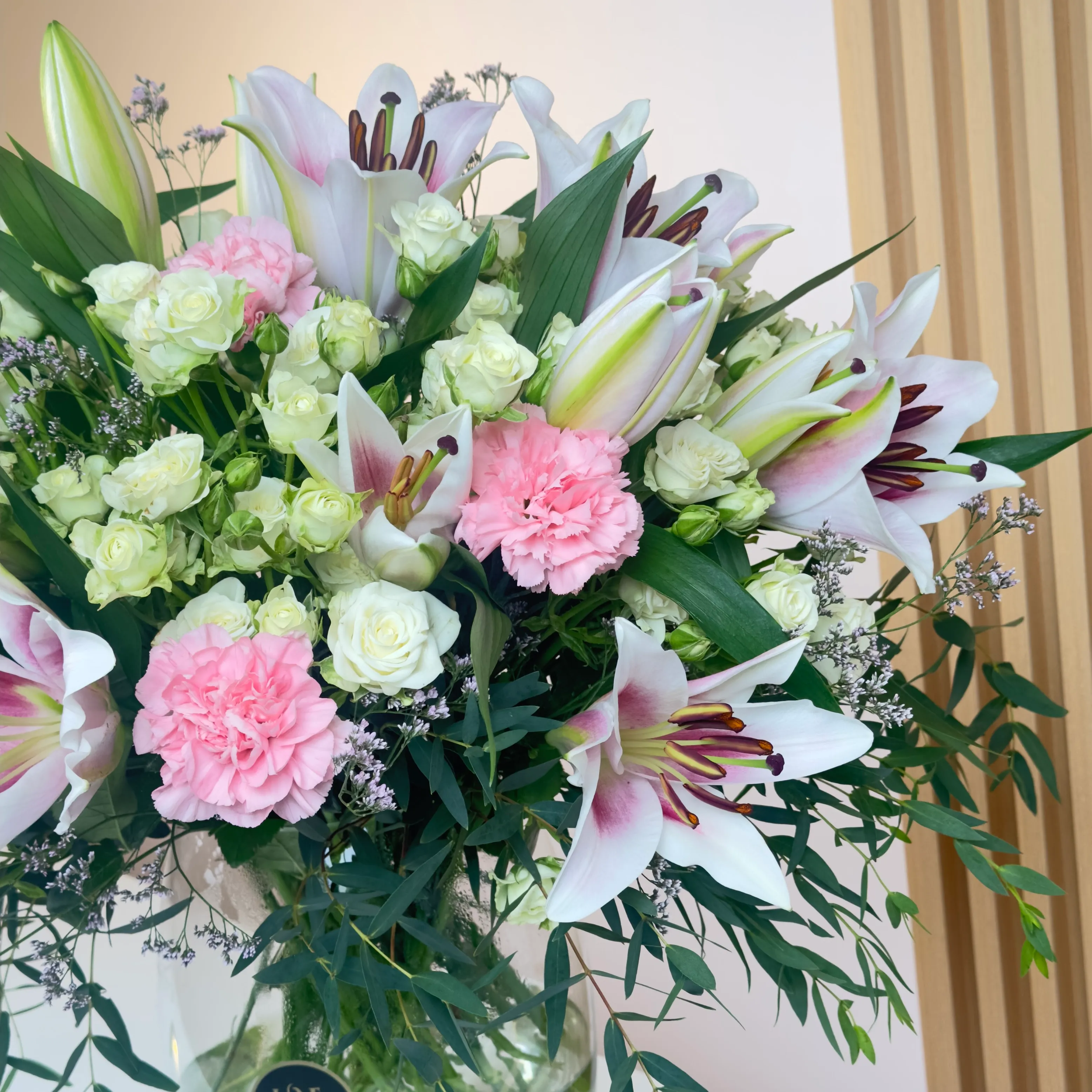 An arrangement of 4 pink lilies, 10 pink carnations, and 10 white spray roses with green fillers in a glass vase