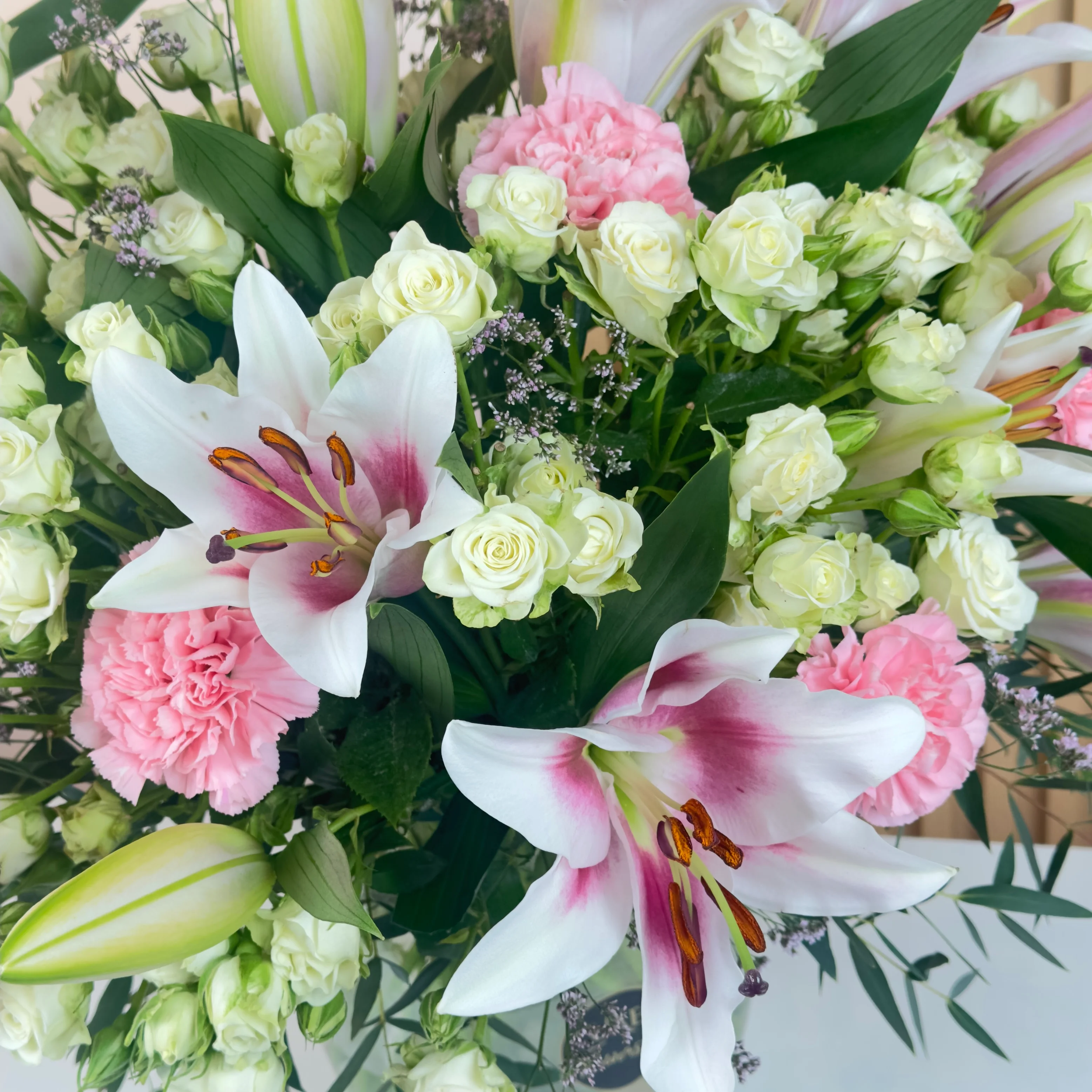 An arrangement of 4 pink lilies, 10 pink carnations, and 10 white spray roses with green fillers in a glass vase