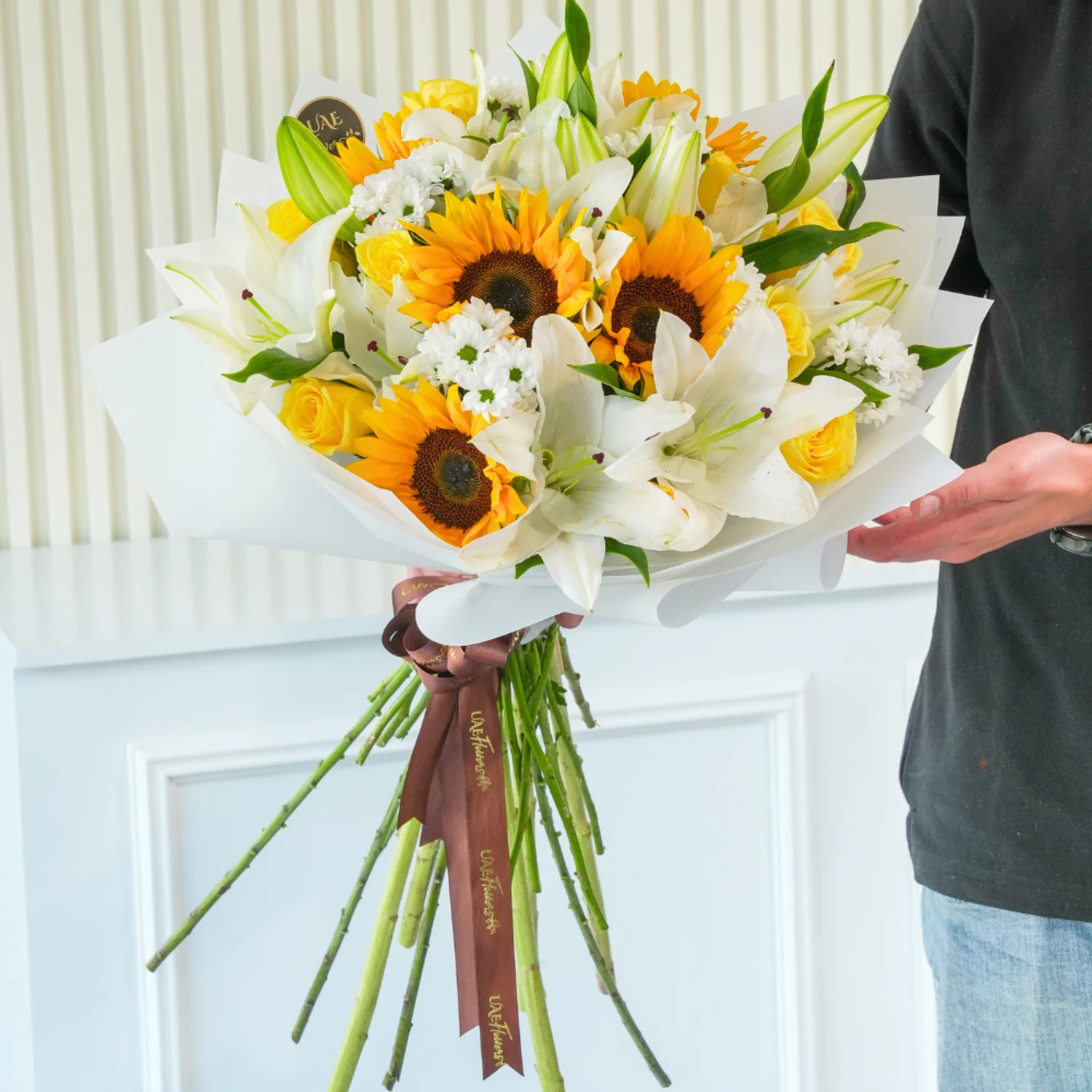 Bright bouquet of sunflowers, white lilies, yellow roses, and white filler flowers wrapped in elegant white paper with a brown satin ribbon – joyful floral gift by UAE Flowers.