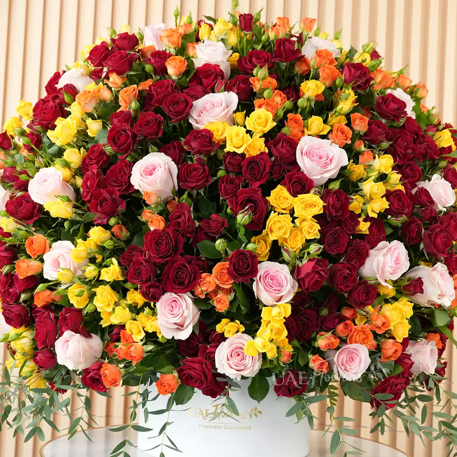 A half-round floral arrangement featuring 30 pink roses and 50 red, orange, and yellow spray roses, along with green fillers and eucalyptus leaves, presented in a white round box