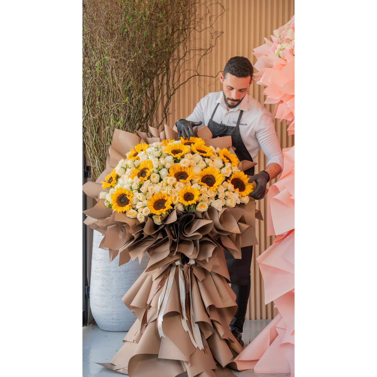 Tall luxury floral stand with 15 sunflowers, 50 spray roses, and 50 premium roses, wrapped in brown draping with satin ribbons – elegant event display by UAE Flowers.