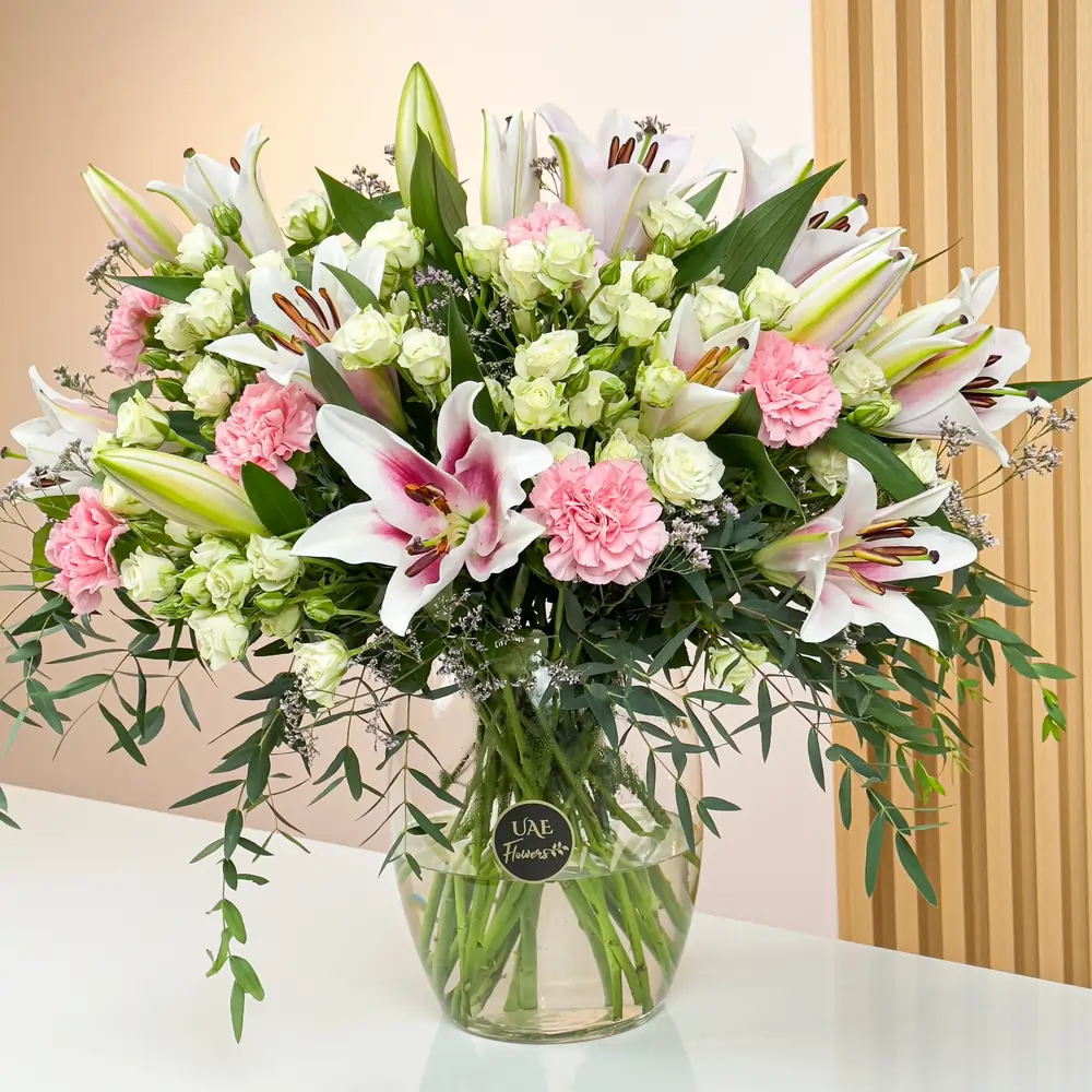 An arrangement of 4 pink lilies, 10 pink carnations, and 10 white spray roses with green fillers in a glass vase