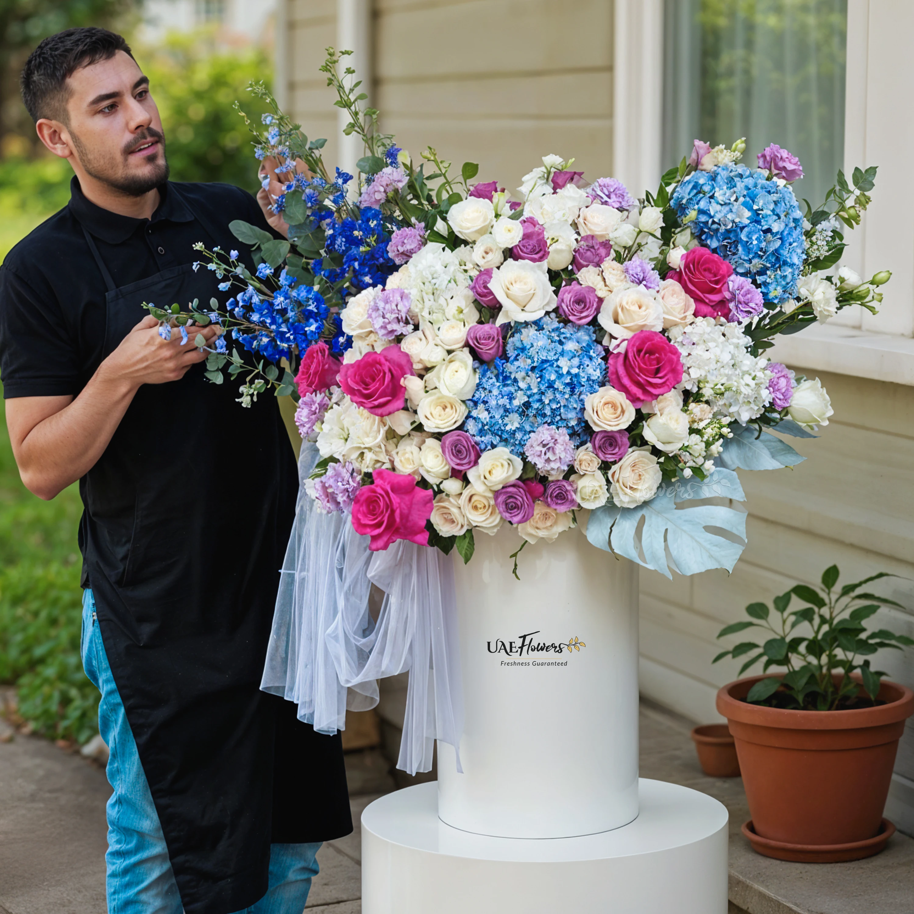 An arrangement of blue hydrangeas, white hydrangeas, purple roses, white roses, purple lisianthus, white spray roses, blue delphinium, fushia roses with green fillers and blue net in a tall round box.