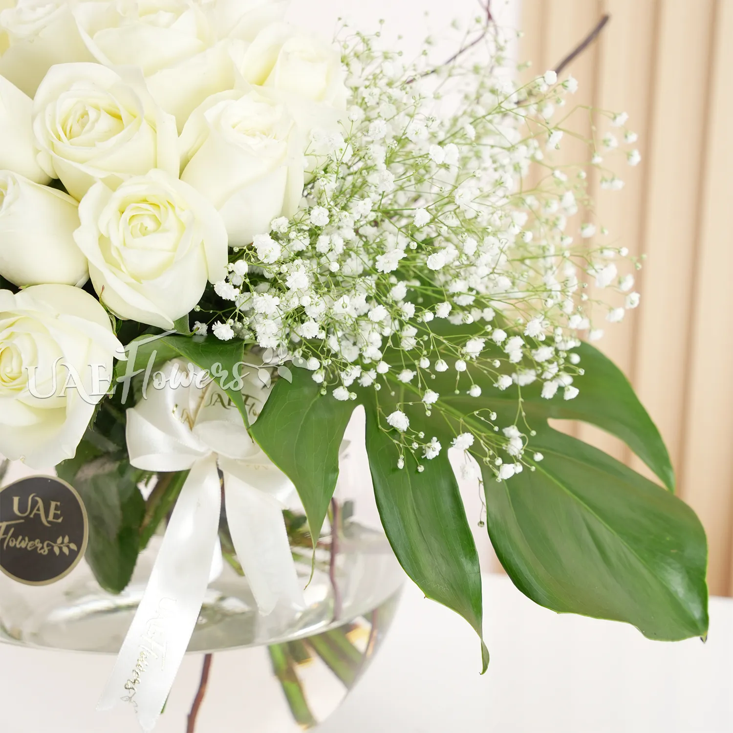 Round glass vase with 20 white roses, baby's breath, and green fillers, elegantly arranged.